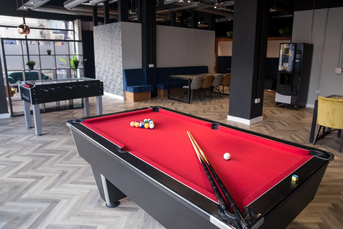 A recreation room with a red pool table, cues, a foosball table, and seating area on a herringbone-patterned floor.