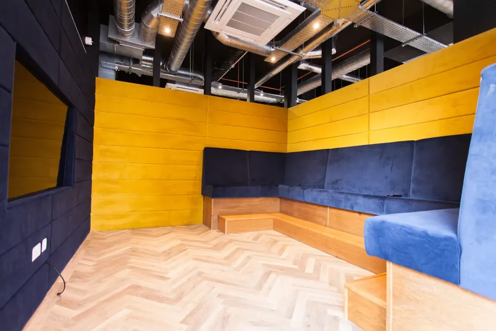 Modern room with yellow and dark blue walls, blue cushioned seating, wooden flooring, and exposed ceiling ducts.