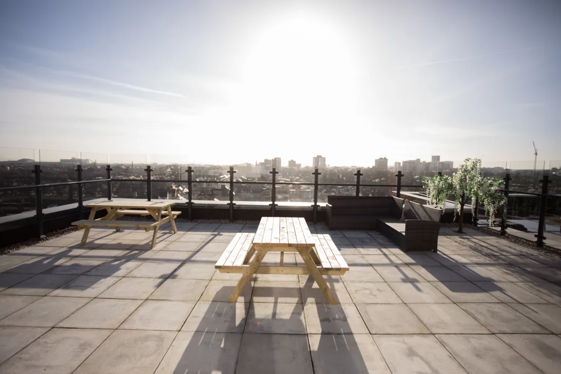 Sunny rooftop terrace with wooden picnic tables, planters, and a city skyline view in the background.