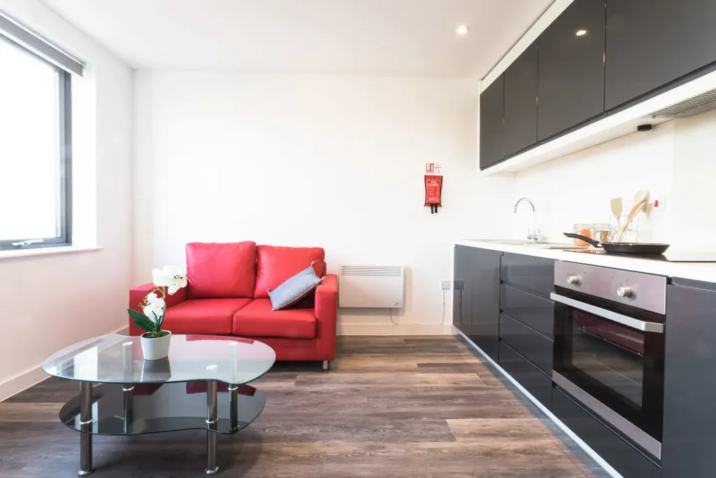 Modern kitchen and living area with black cabinets, red sofa, glass coffee table, and wood flooring. Bright window light illuminates the space.