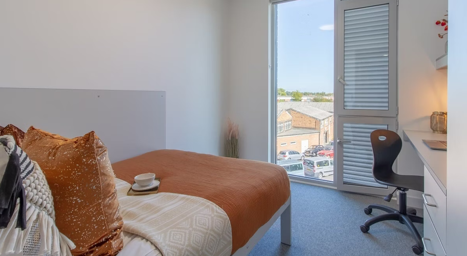 Bright bedroom with a neatly made bed, orange throw, pillows, and a cup. Desk with chair by a window overlooking buildings.