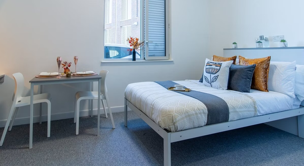 Modern bedroom with a neatly made bed, decorative pillows, a small dining table with two chairs, and a window with blinds.