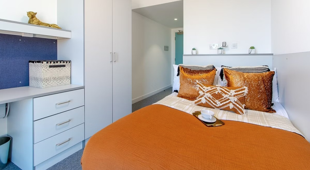 Cozy bedroom with a bed featuring orange pillows and blanket, white cabinets, a desk, and a decorative box. A cup sits on a tray on the bed.