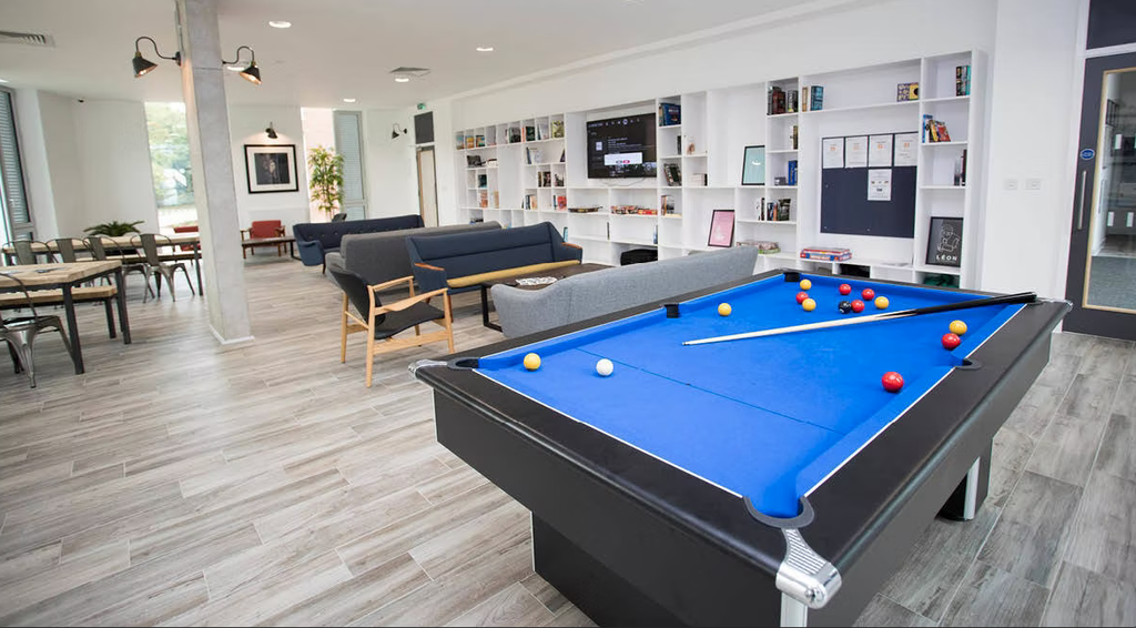 Modern lounge area with a blue pool table, sofas, a TV, bookshelves, and large windows providing natural light.