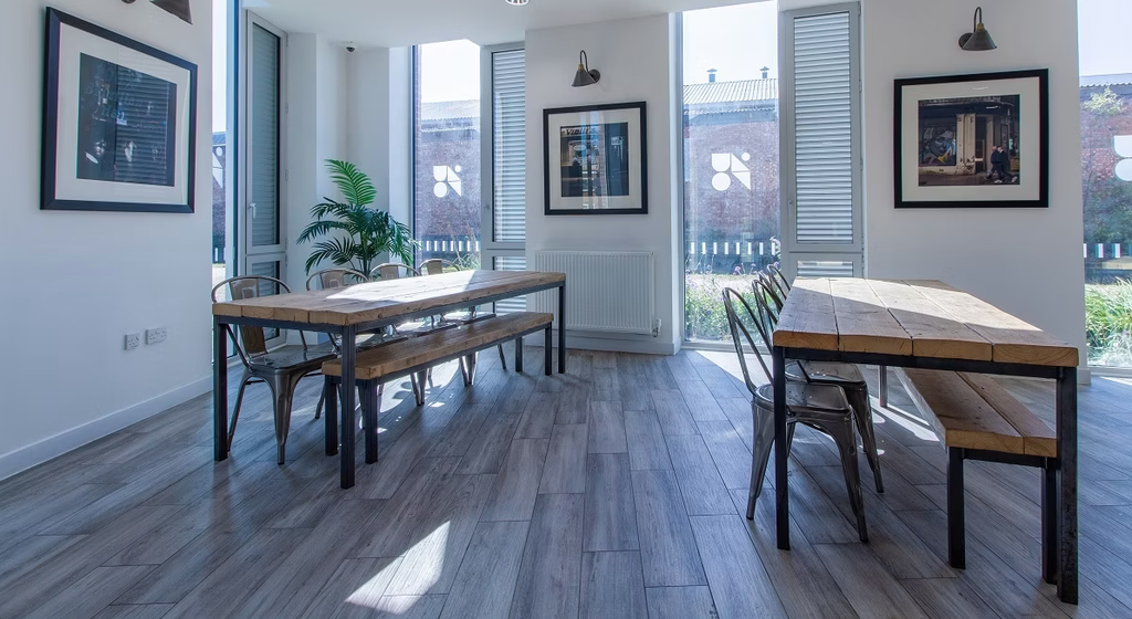 Bright room with two wooden tables, metal chairs, framed art on walls, large windows, and a potted plant, creating a modern, airy atmosphere.