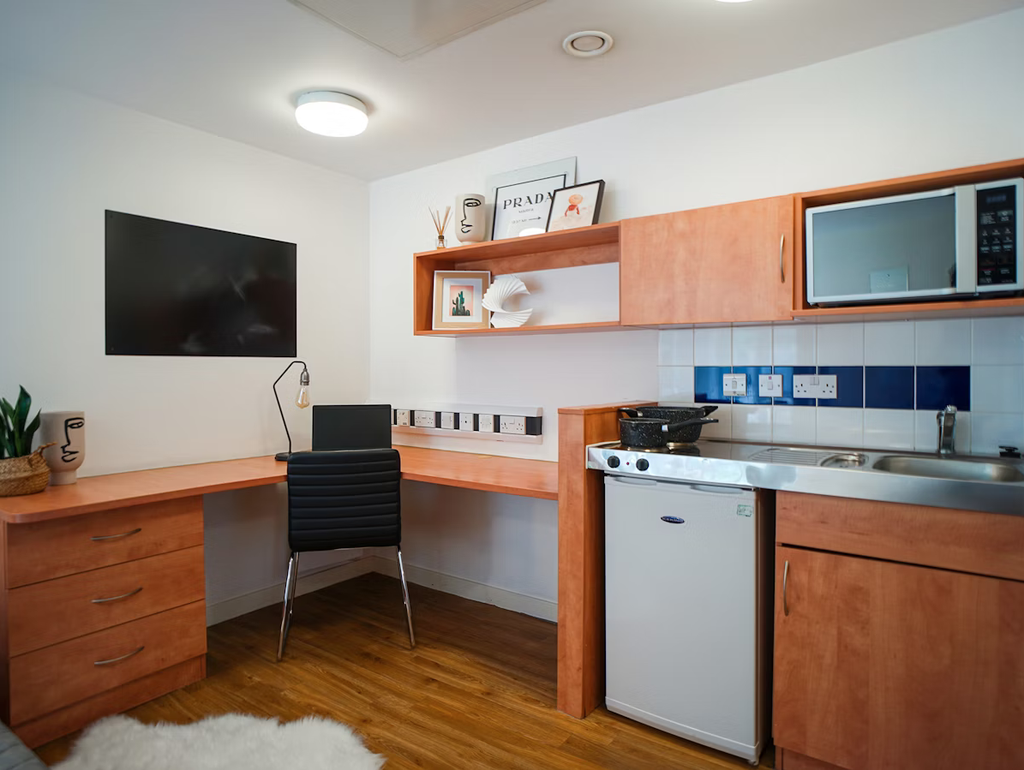 Modern dorm room with wooden desk, black chair, wall-mounted TV, shelves with decor, mini-fridge, microwave, and small sink area.