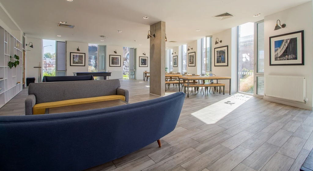 Modern lounge with blue sofas, wooden tables, and framed art on walls. Large windows let in natural light, creating an airy atmosphere.