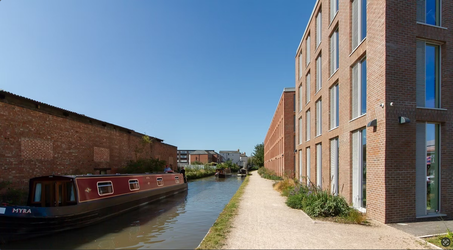 A narrowboat on a canal beside a brick building under a clear blue sky, with a path and another building on the opposite side.