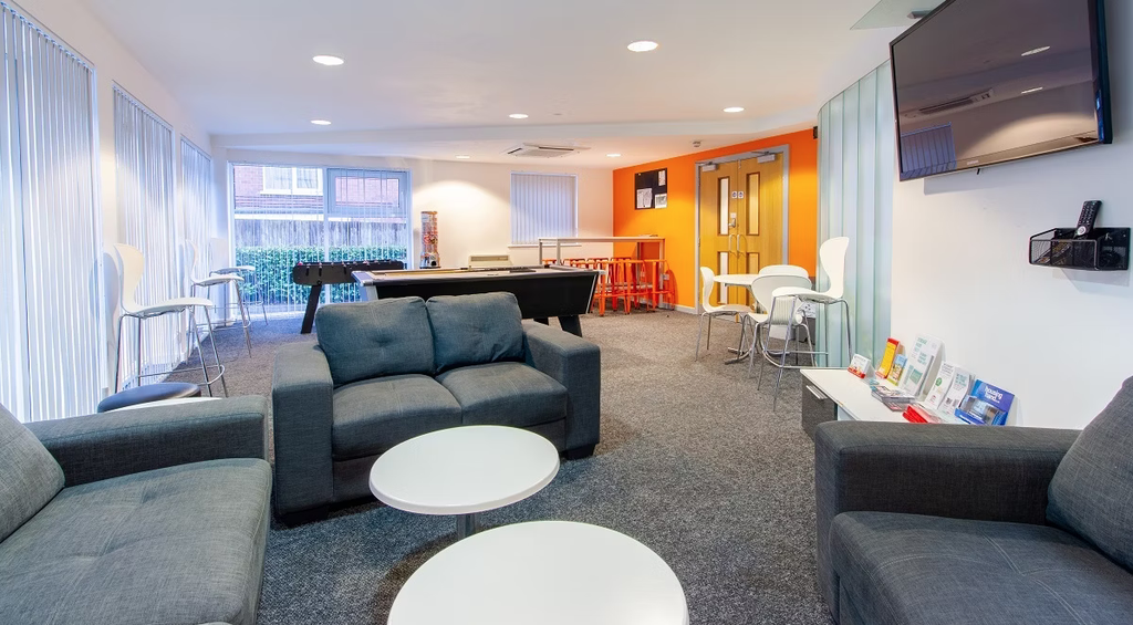 Modern lounge with gray sofas, round white tables, a foosball table, bar stools, wall-mounted TV, and brochures on a stand, lit by natural light.