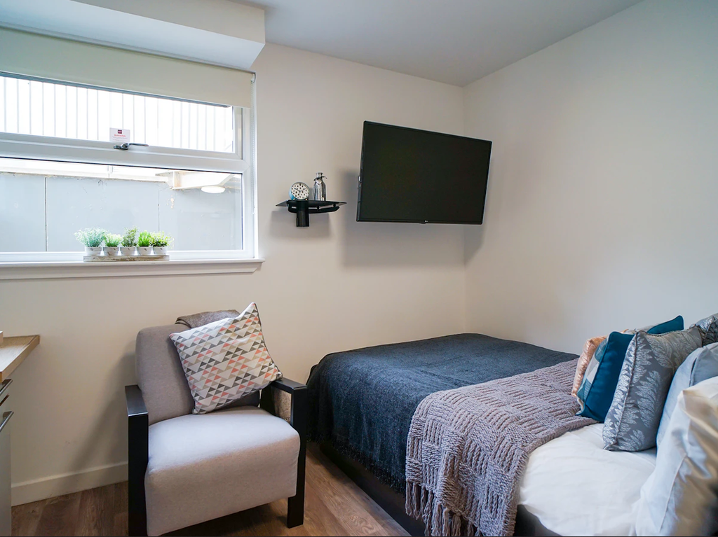 A cozy bedroom with a bed, pillows, a wall-mounted TV, an armchair, a small shelf, and a window with potted plants on the sill.