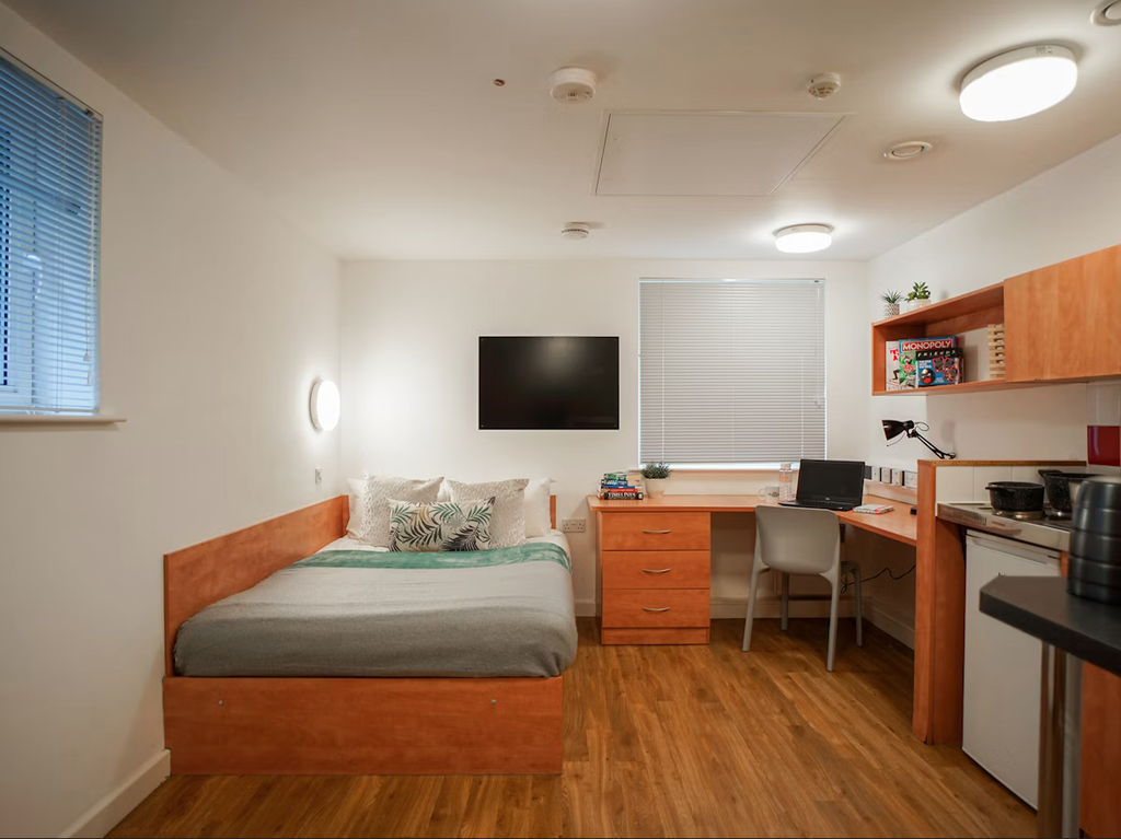 A tidy dorm room with a bed, desk, chair, and kitchenette. The room has wooden flooring and a wall-mounted TV.