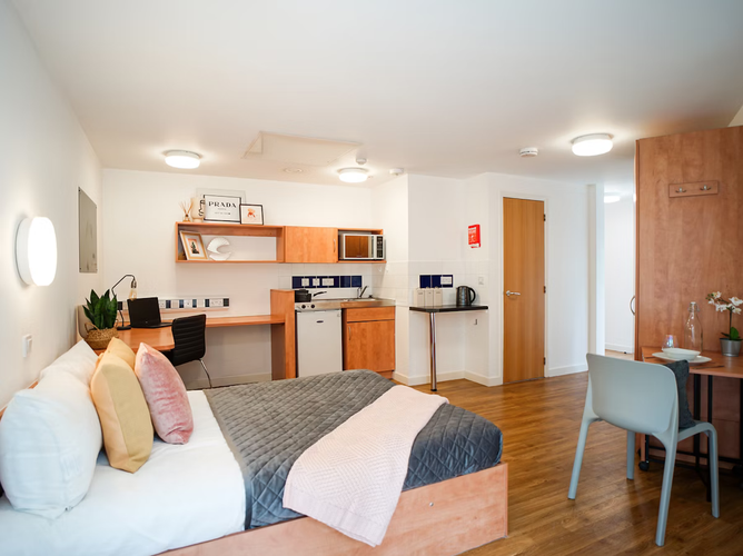 Modern studio apartment with a bed, desk, kitchenette, and dining area. Warm wood tones, decorative shelves, and cozy lighting create a welcoming atmosphere.
