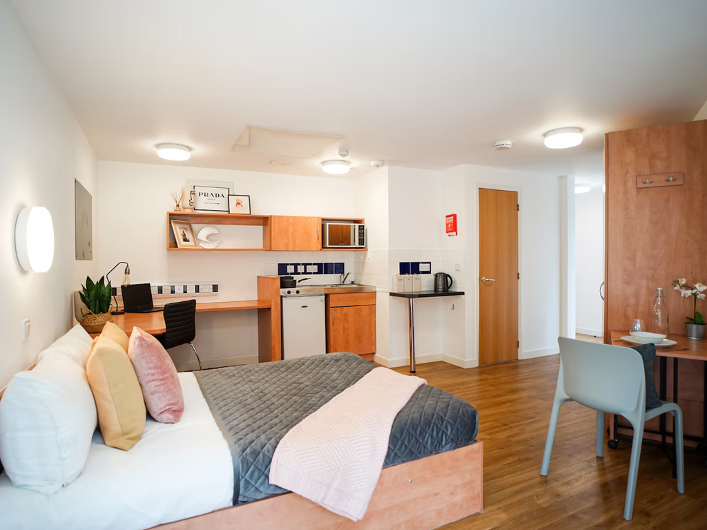 Modern studio apartment with a bed, desk, kitchenette, and dining area. Warm wood tones, decorative shelves, and cozy lighting create a welcoming atmosphere.