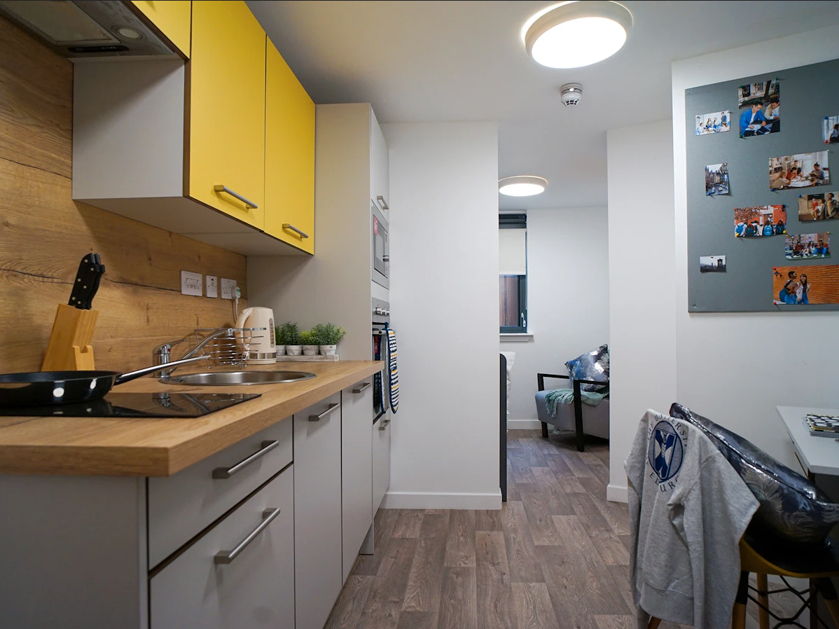 Modern kitchen with yellow cabinets, wooden countertop, and appliances. A cozy room with a bed and photo-decorated wall is visible in the background.