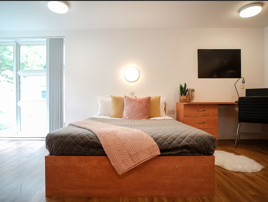 Modern bedroom with a neatly made bed, pink and gray bedding, wall-mounted TV, desk with a chair, and a window with blinds.