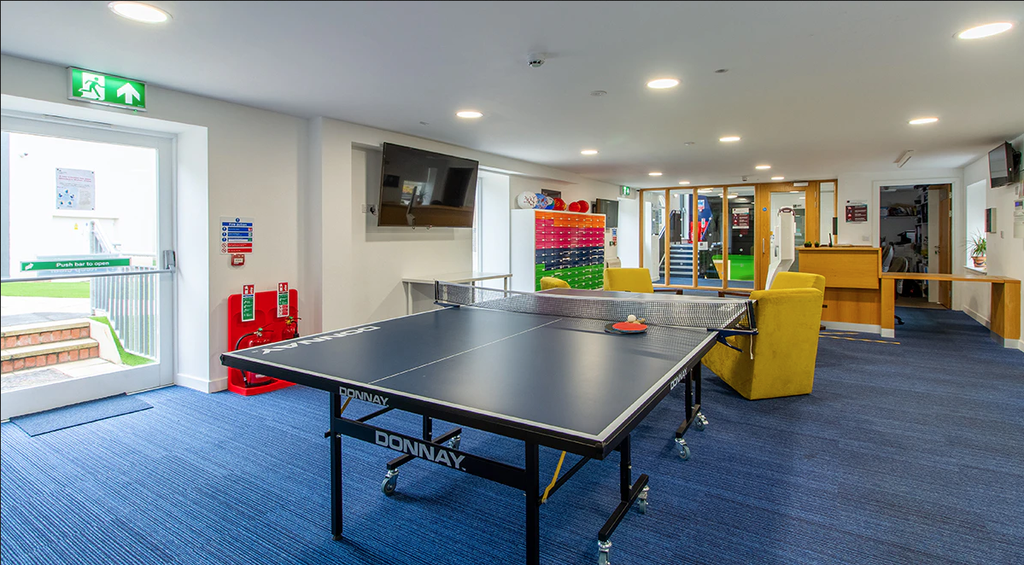 Recreational room with a ping pong table, yellow chairs, wall-mounted TV, and emergency exit signs, featuring blue carpeted flooring.