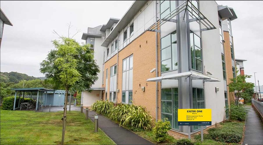 Modern three-story building with large windows, surrounded by greenery and bicycle parking, featuring a yellow sign labeled "Exeter One."