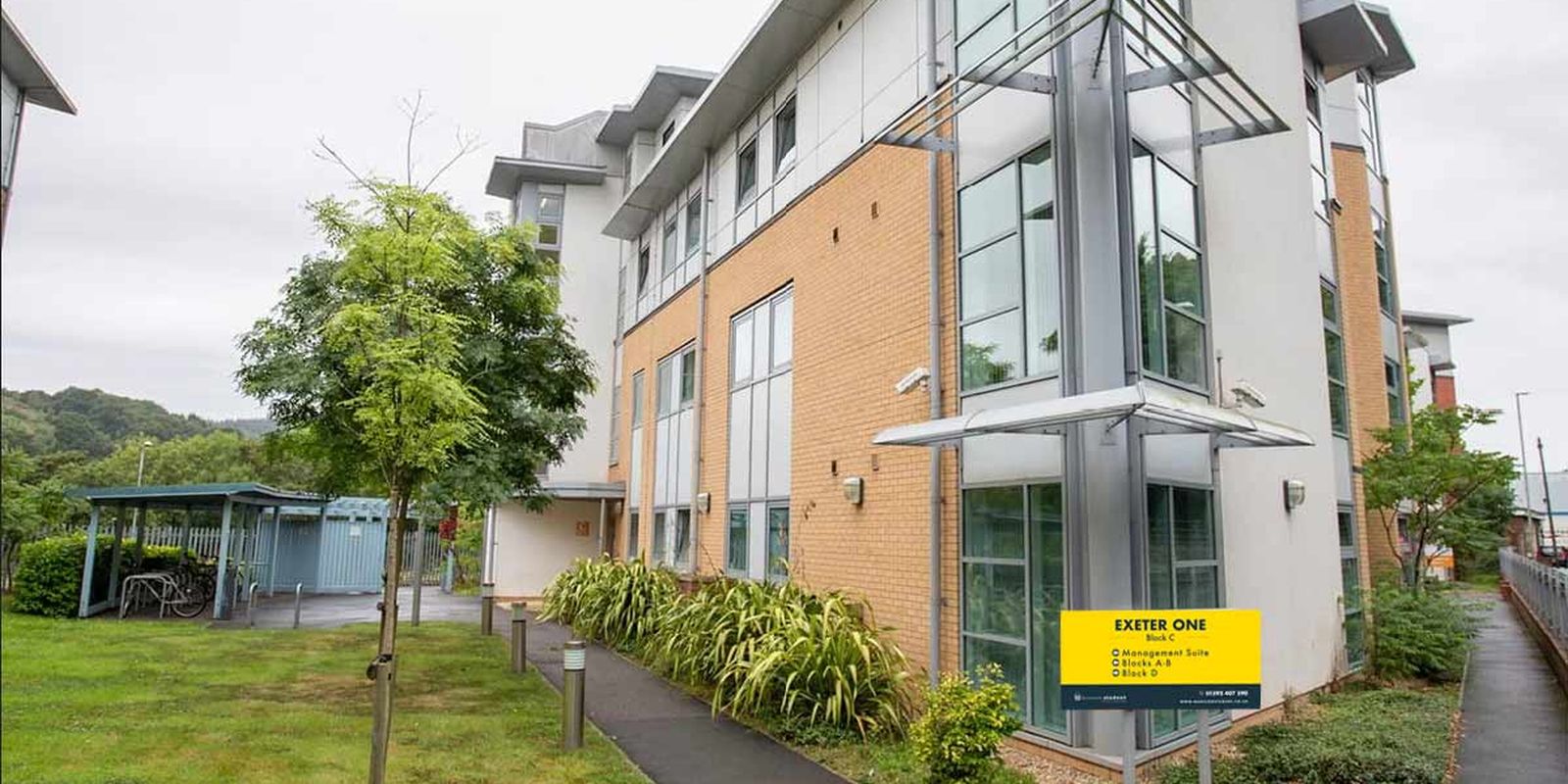 Modern three-story building with large windows, surrounded by greenery and bicycle parking, featuring a yellow sign labeled "Exeter One."