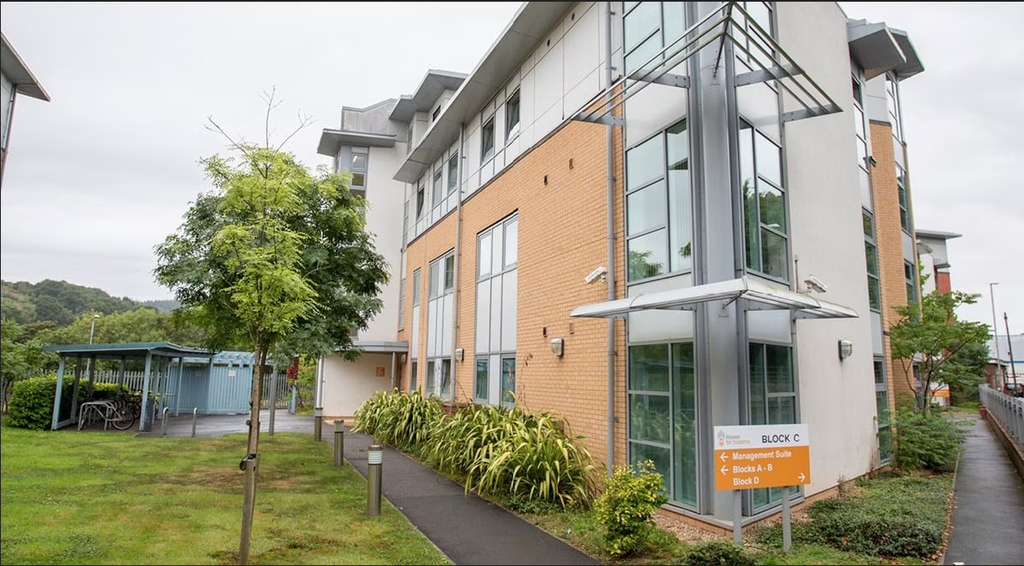 Modern three-story building with large windows, surrounded by greenery. A sign indicating directions is in the foreground.