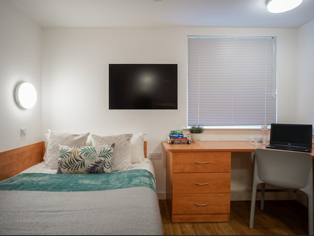 A tidy bedroom with a bed, wall-mounted TV, wooden desk with books and a laptop, chair, and a window with blinds.