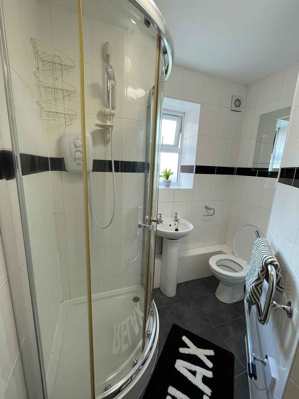 A small bathroom with a curved glass shower, sink, toilet, and a window. A black rug says "RELAX." White tiles and a striped towel are visible.