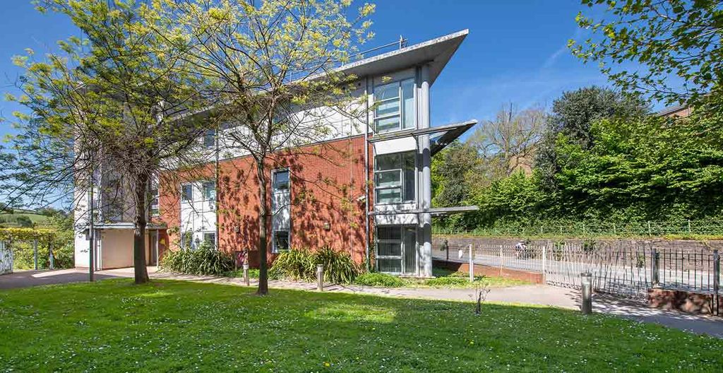 Modern building with red brick facade and large glass windows, surrounded by trees and greenery on a sunny day.