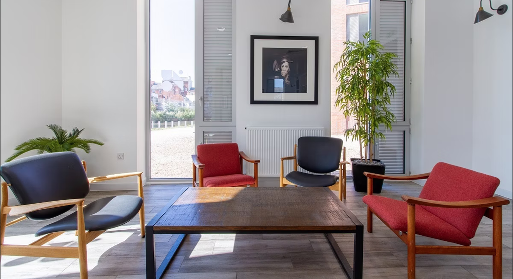 Modern office lounge with wooden chairs, red cushions, a central table, potted plants, and a large window with a city view.
