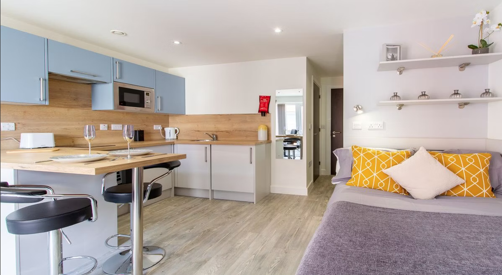 Modern studio apartment with a kitchenette, breakfast bar, and cozy seating area. Features blue cabinets, wood accents, and decorative pillows.