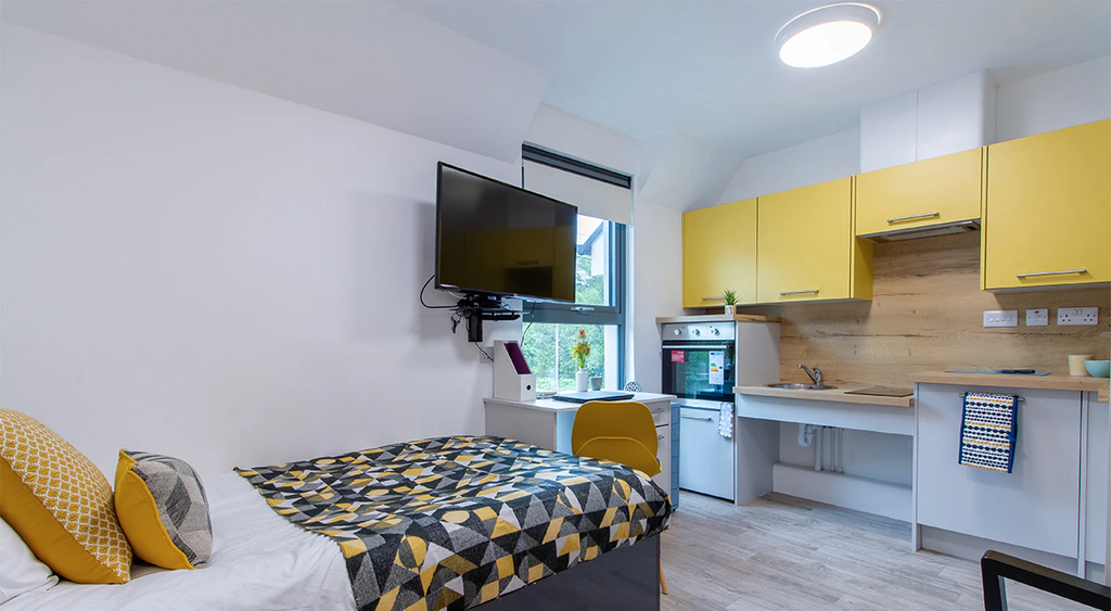 Modern studio apartment with a bed, wall-mounted TV, desk, yellow cabinets, and a small kitchen area. Bright and neatly arranged.