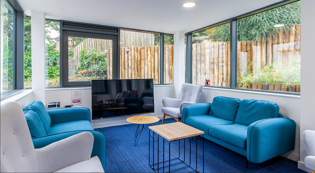 A bright, modern living room with blue sofas, a wooden coffee table, and a large TV. Large windows offer a view of a fenced garden area.