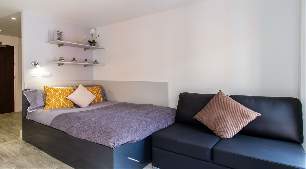 Modern bedroom with a bed featuring gray and yellow pillows, a black sofa, wall shelves, and light wood flooring.