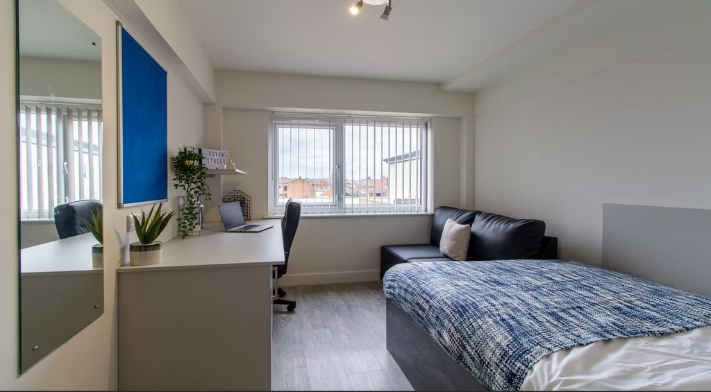 Modern bedroom with a single bed, blue blanket, desk with a computer, potted plants, and large windows with vertical blinds.
