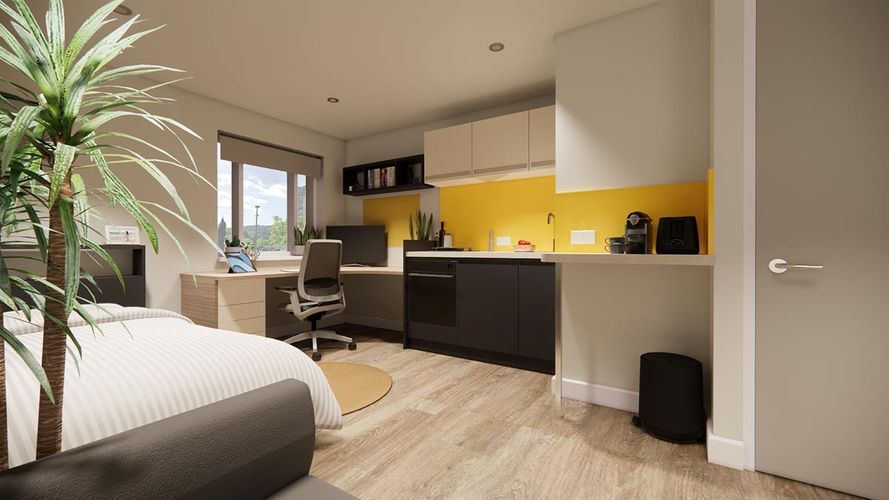 Bright modern studio with bed, large potted plant, L-shaped desk and office chair by window, kitchenette with yellow backsplash and wood floors.