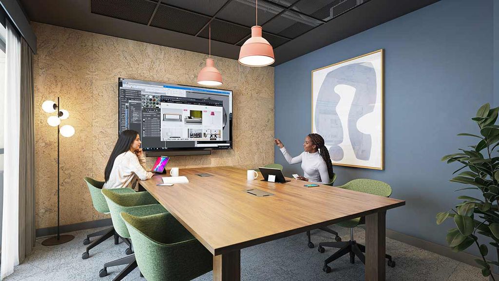 Two women in a modern conference room at a wooden table, one pointing to a large wall monitor displaying web designs, green chairs and plants.