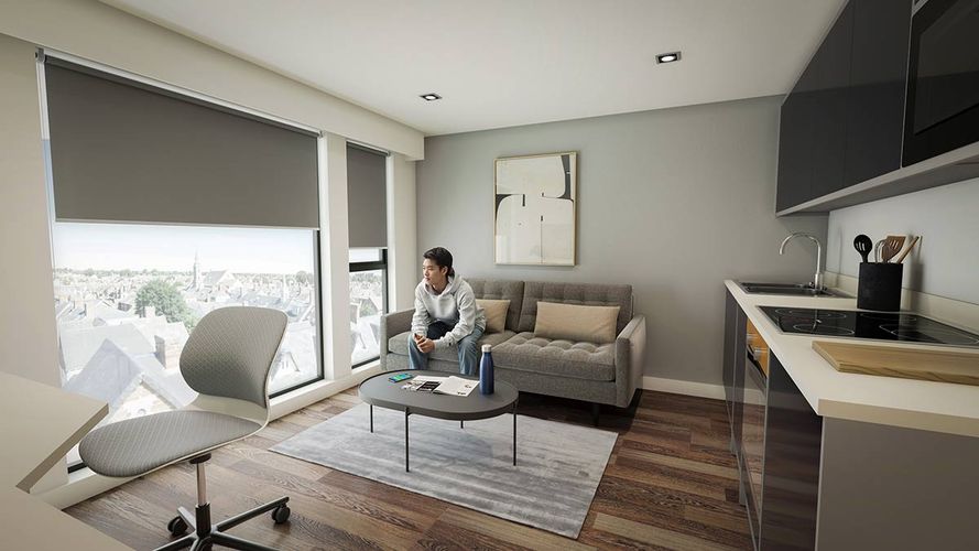Modern studio apartment interior with sofa, coffee table and person seated by large windows overlooking city skyline; kitchenette to right.