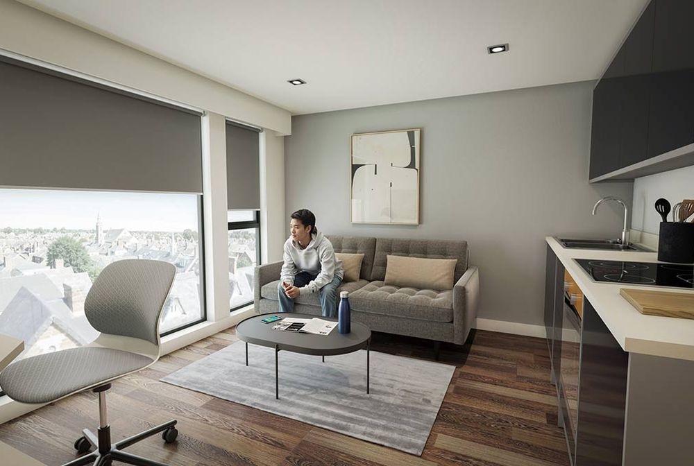 Modern studio apartment interior with sofa, coffee table and person seated by large windows overlooking city skyline; kitchenette to right.