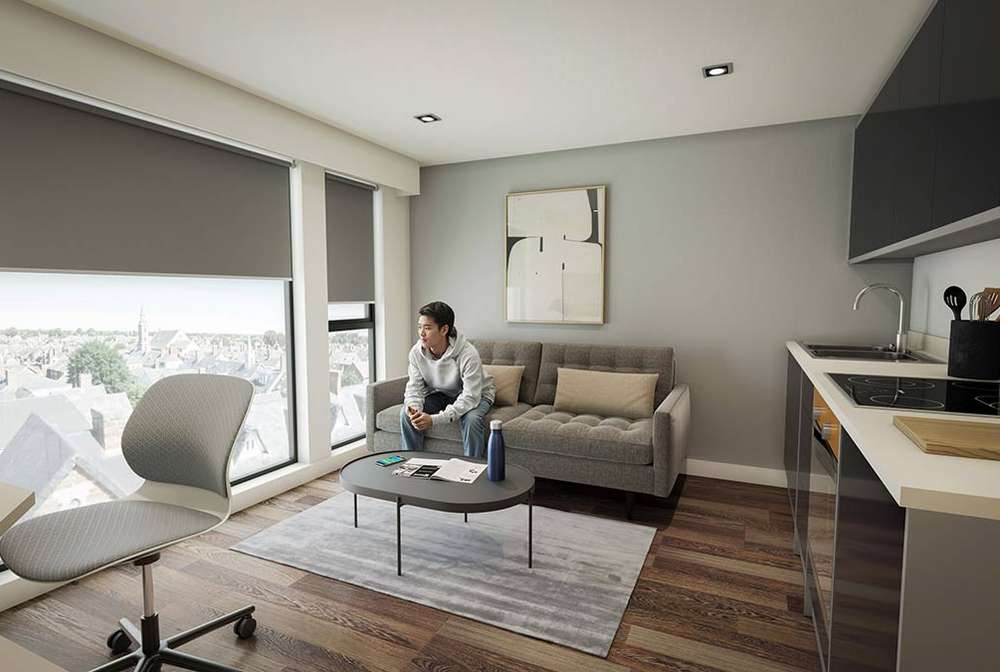 Modern studio apartment interior with sofa, coffee table and person seated by large windows overlooking city skyline; kitchenette to right.