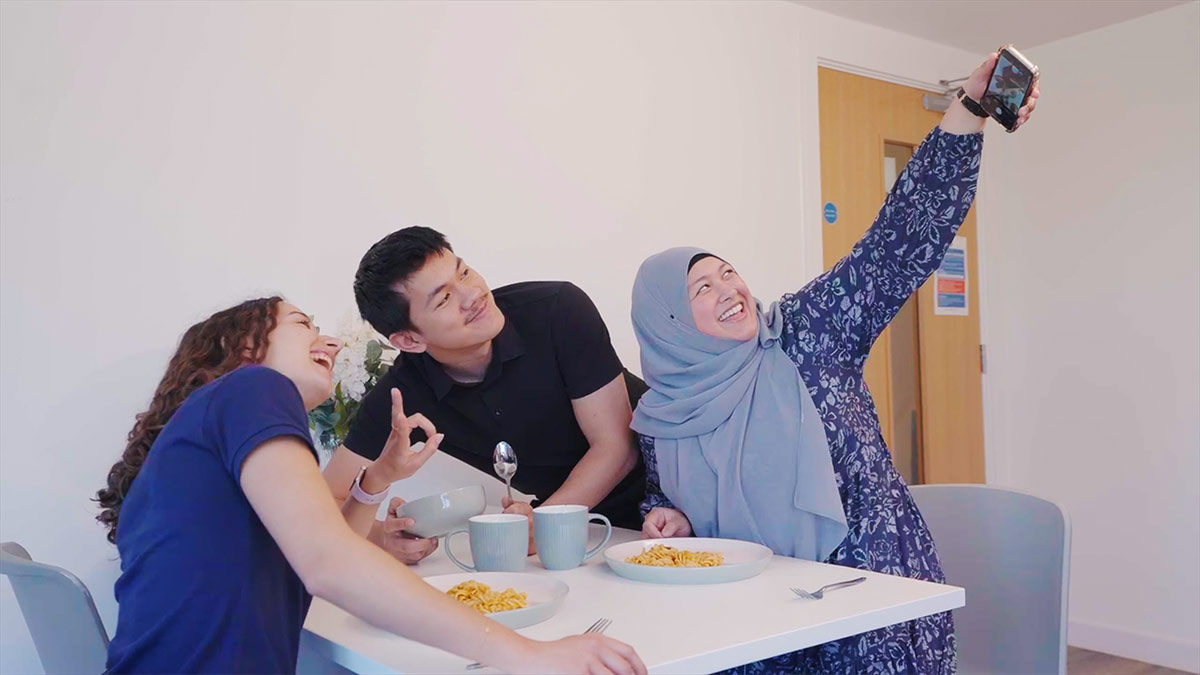 Three friends smile while taking a selfie at a table with plates of pasta and cups, enjoying a meal together in a well-lit room.