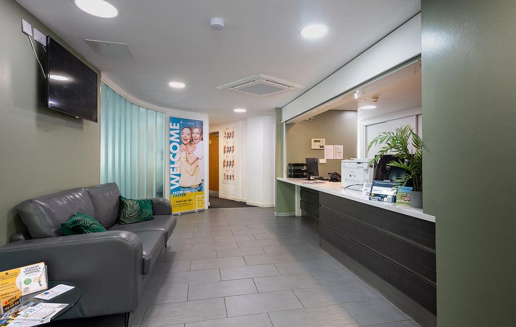 Modern office reception with a sofa, welcome banner, counter with a printer, potted plant, and brochures, under bright lighting.