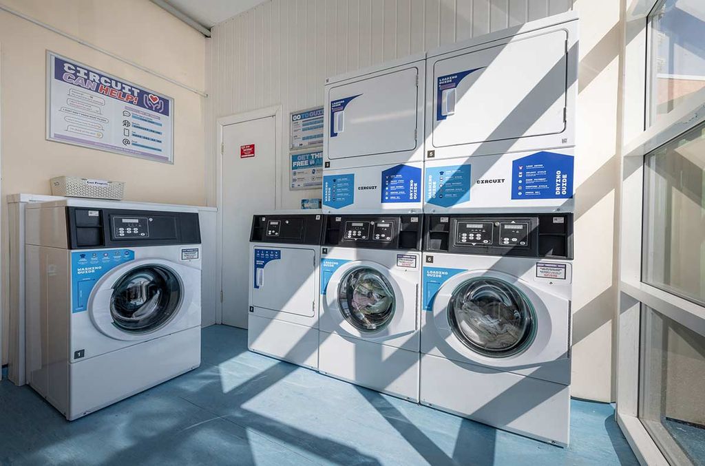 A bright laundromat with stacked washers and dryers, blue floor, and sunlight streaming in from large windows, creating shadows on the machines.