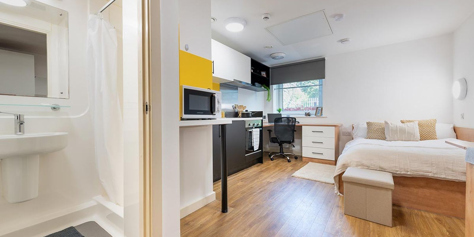 A modern studio apartment with a bed, desk, kitchenette, and a bathroom. Wooden flooring and natural light create a cozy, functional space.