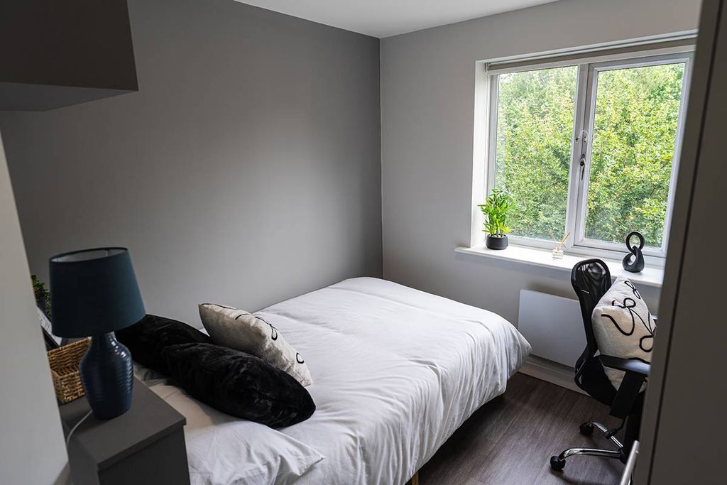 Bright bedroom with a neatly made bed, black and white pillows, a black lamp, desk with a chair, potted plants, and a window with greenery outside.