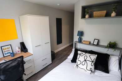 Modern bedroom with a white bed, black and white pillows, desk with chair, yellow pinboard, wardrobe, shelves with plants, and a blue lamp.