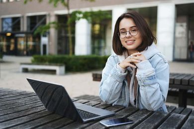 A person wearing glasses and a denim jacket sits at an outdoor table with a laptop and smartphone, smiling at the camera.