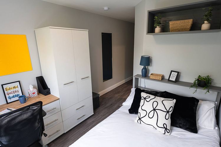 A modern bedroom with a white wardrobe, desk, chair, bed, decorative pillows, lamp, and wall shelf with plants and a basket.