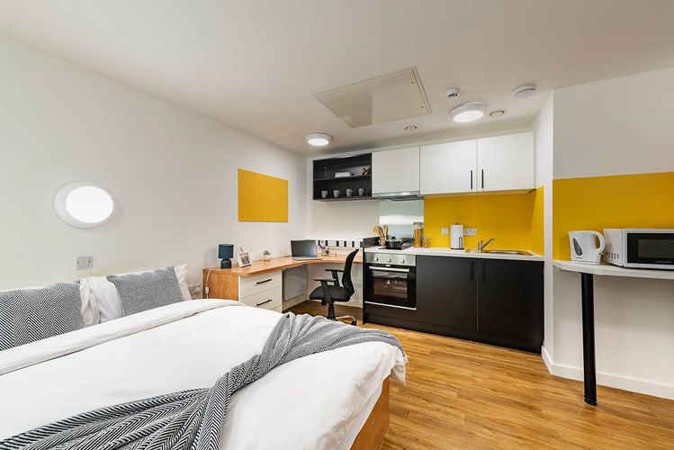 Modern studio apartment with a bed, desk, and kitchen area. Features wood flooring, yellow walls, and compact appliances.
