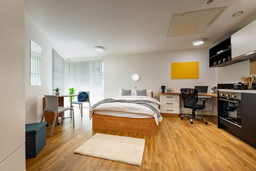 Modern studio apartment with a bed, desk, kitchenette, and dining area on wooden flooring, featuring natural light and simple decor.