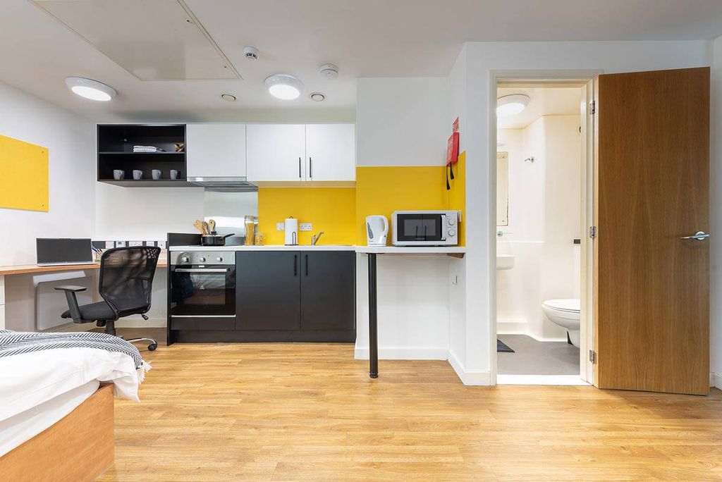Modern studio apartment with a workspace, kitchen, bed, and open door to the bathroom. Bright yellow accents and wooden flooring.