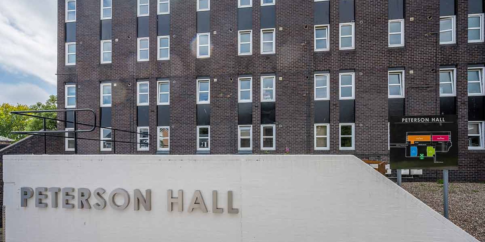 A brick building named Peterson Hall with a white sign in front and a map beside the entrance, under a partially cloudy sky.