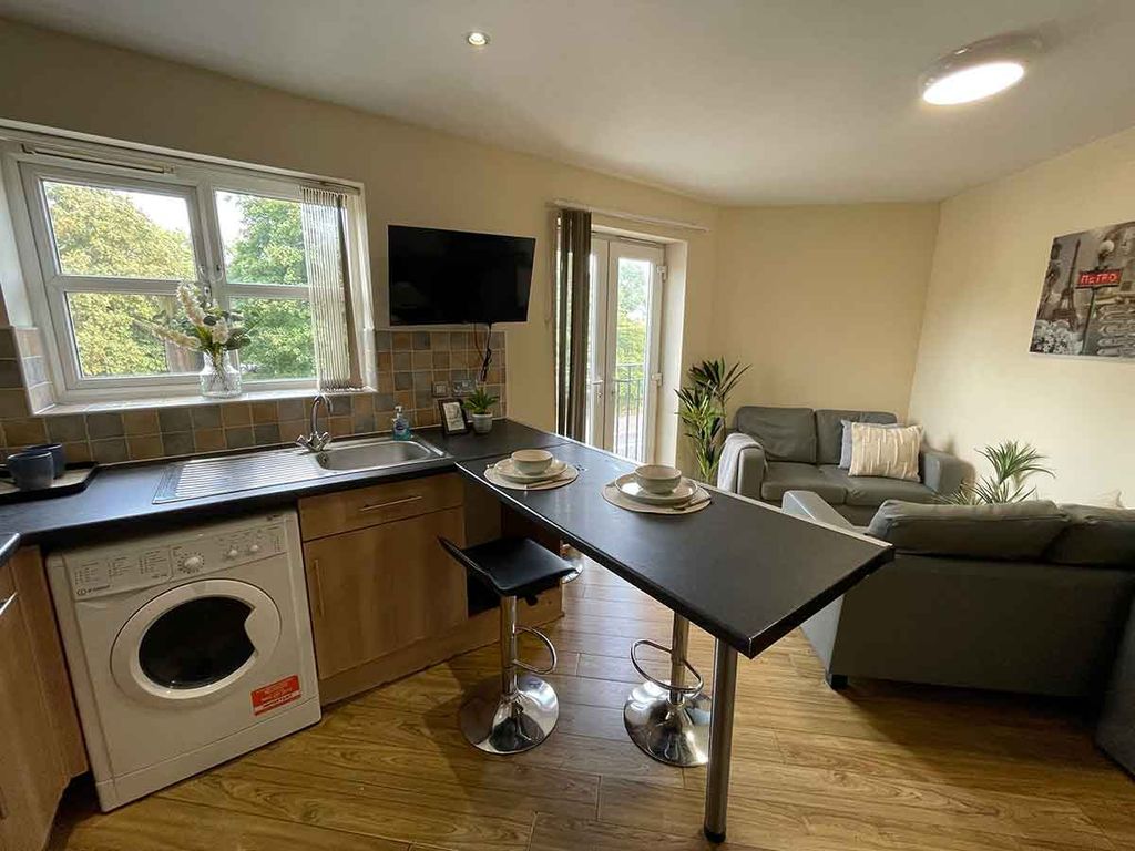 Modern open-plan kitchen and living area with wooden floors, bar seating, a sofa, wall-mounted TV, and large windows overlooking greenery.